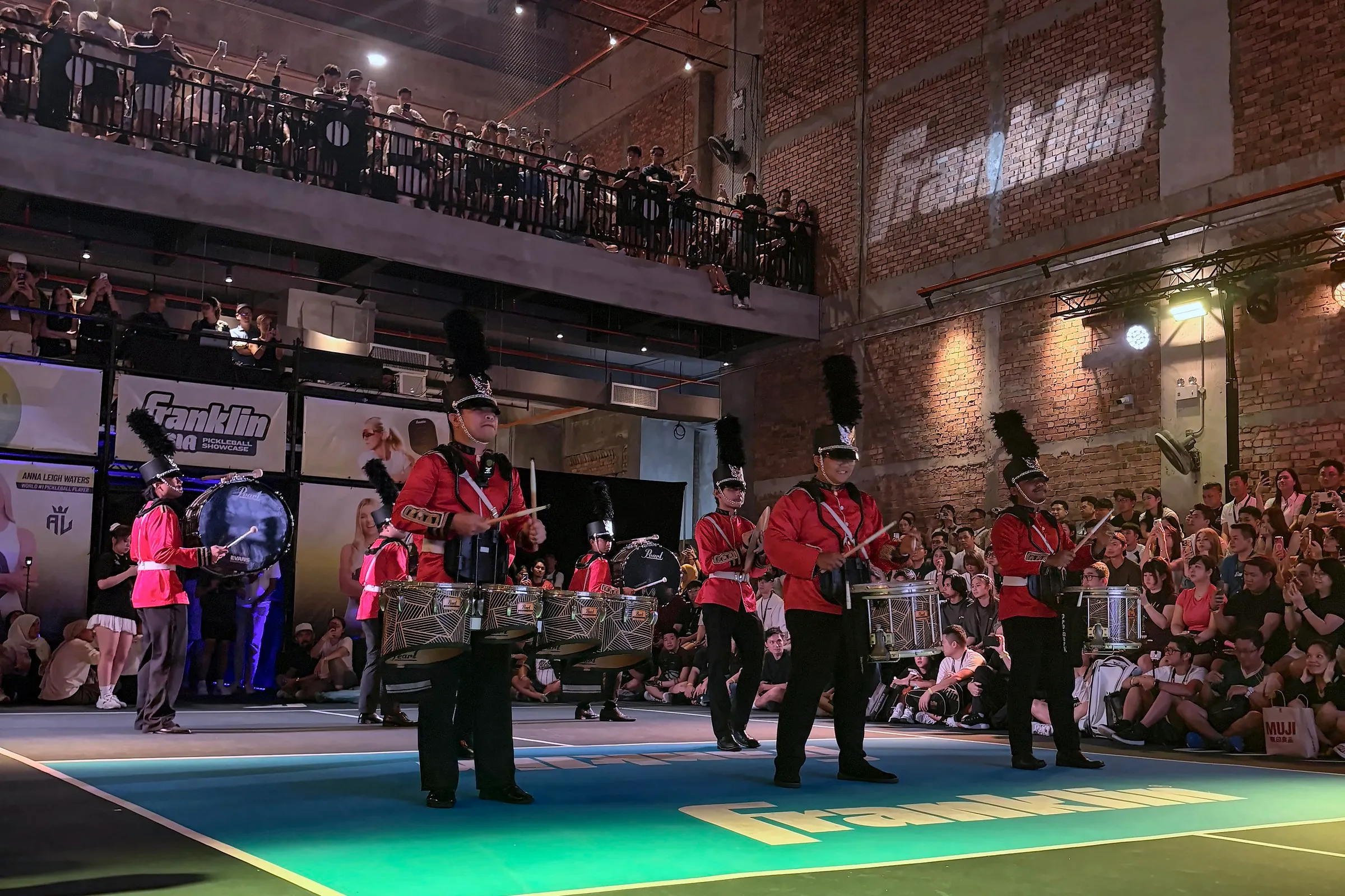 A marching drum band in red tunics plays on court at the start of the showcase, with the crowd seated around the edge and in the gallery above