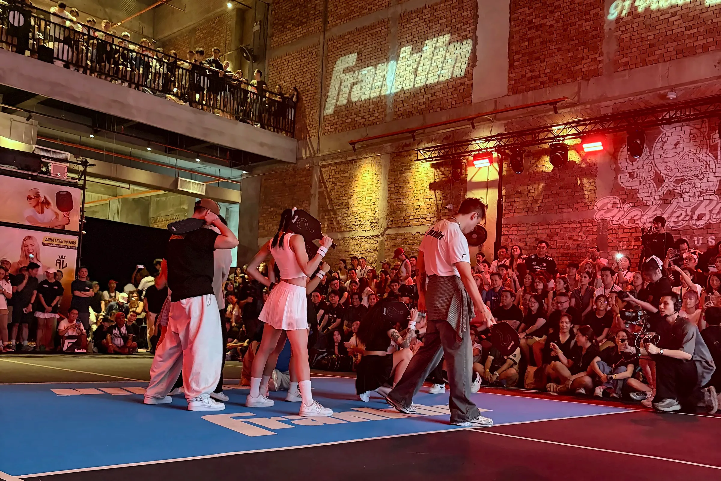 Dancers perform on court under red stage light, with the Franklin and 91 Pickleball names projected on the brick wall behind