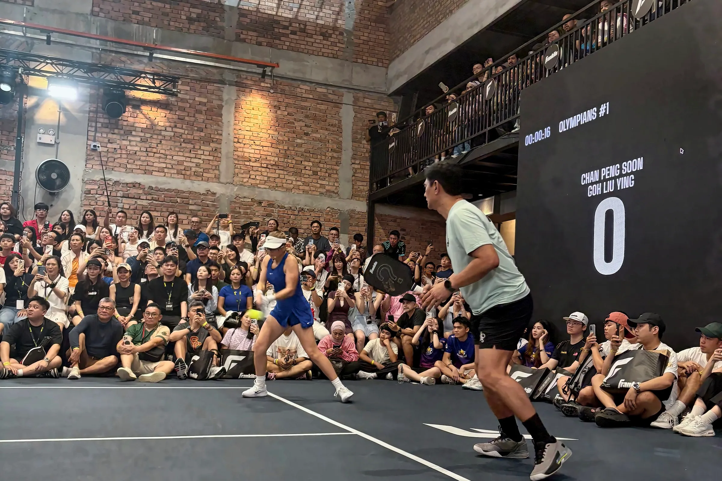 Chan Peng Soon moves forward for a ball, with Anna Leigh Waters on court behind him, the scoreboard reading Olympians and the crowd packed behind