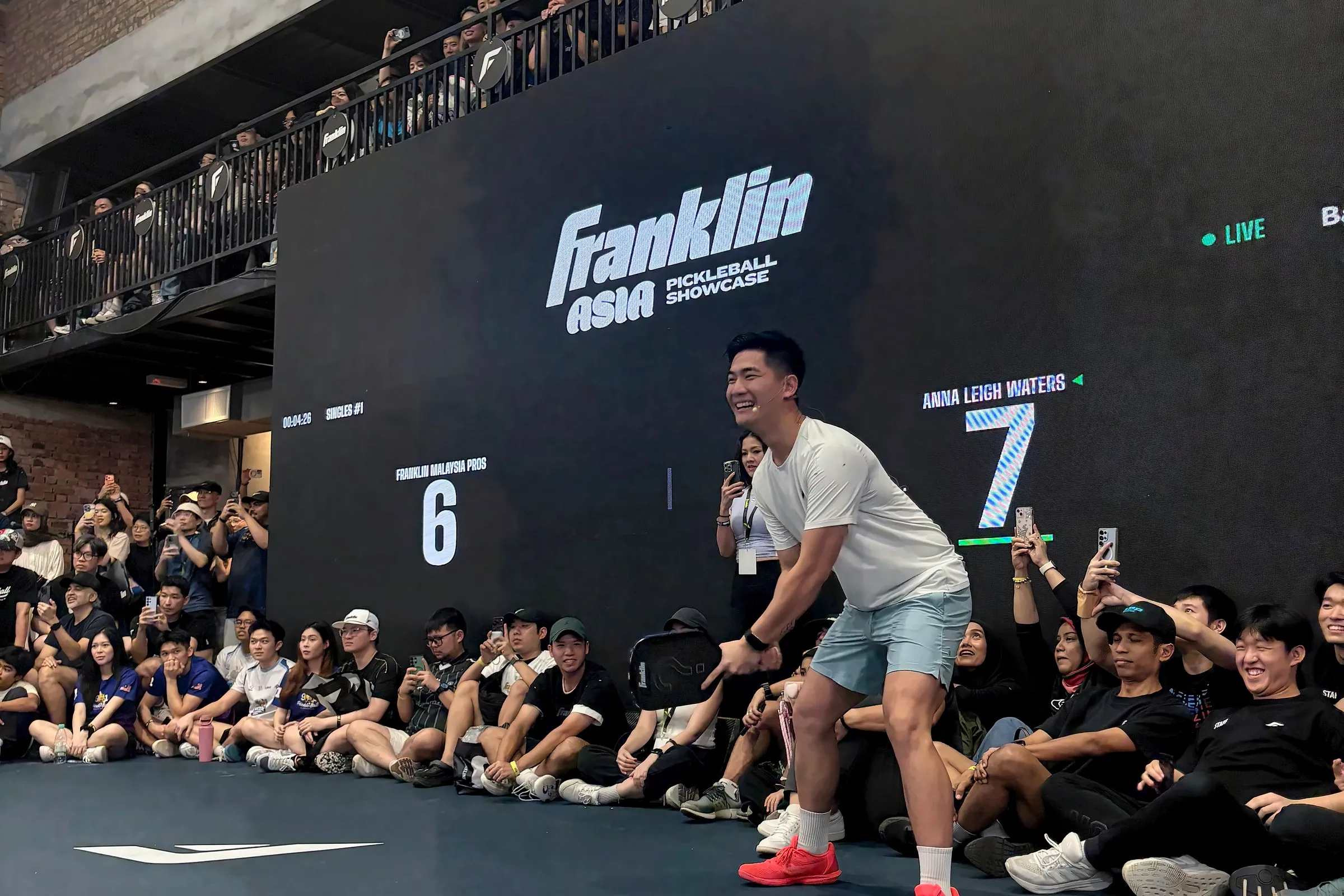 A player in a white T-shirt crouches and grins at the baseline, under a scoreboard reading Franklin Malaysia Pros 6, Anna Leigh Waters 7