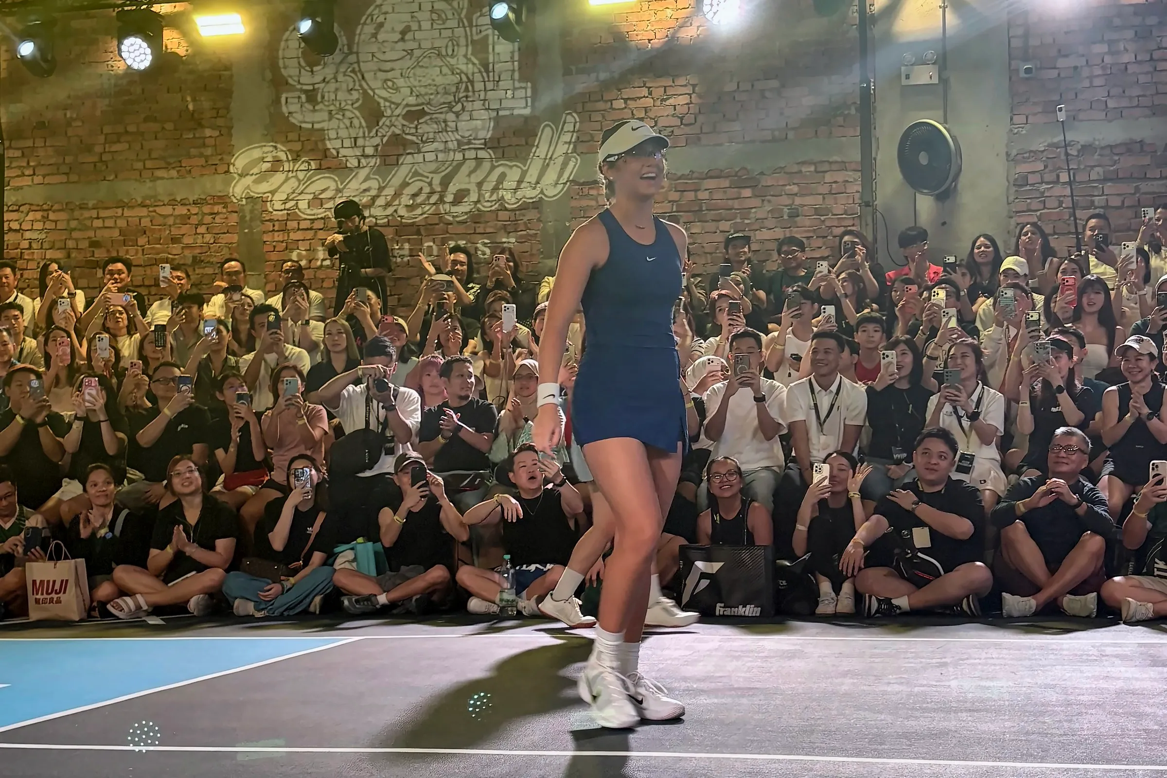 Anna Leigh Waters, in a blue dress and white visor, smiles as she walks along the baseline in front of rows of fans sitting on the floor