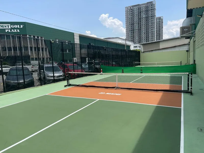 Two green outdoor pickleball courts with orange kitchen zones and fixed nets, inside a black mesh fence next to industrial buildings in Subang Jaya