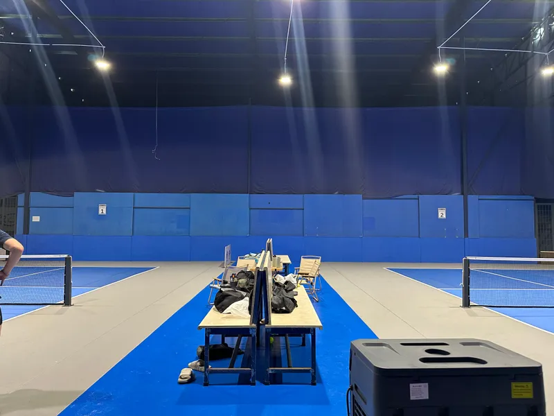 Two indoor pickleball courts with blue and grey surfaces and a central bench area, under bright overhead lights.