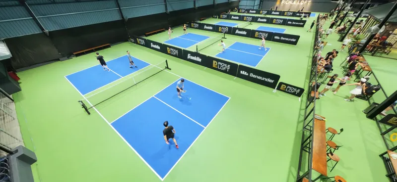 Elevated view of Pickle Loft's indoor hall: blue pickleball courts on green flooring, with small players mid-game and spectators seated along the side.
