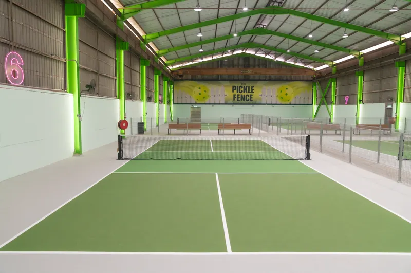 Empty indoor green pickleball court at The Pickle Fence, with a lime-green steel-framed hall, court number 6 on the wall and The Pickle Fence sign at the far end