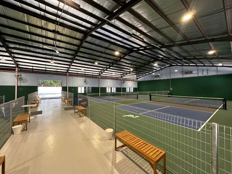 Covered hall at P3 Courts @ Midah with blue-and-green pickleball courts behind wire fencing, and wooden benches along a walkway