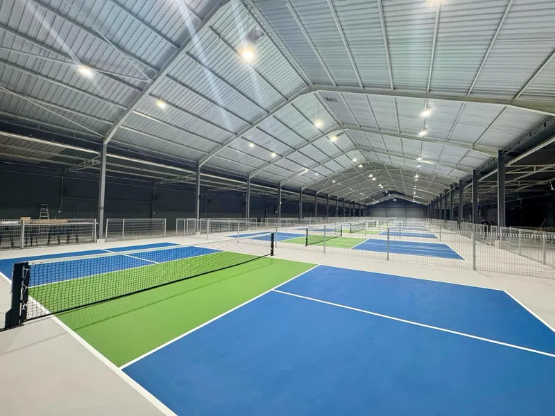 Rows of empty blue-and-green indoor pickleball courts, separated by low mesh fences, under a long steel-roofed hall at PickleTime Subang 7.