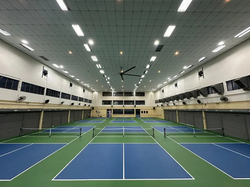 Indoor school hall with blue and green pickleball courts, nets set up, under a high tiled ceiling with a large fan