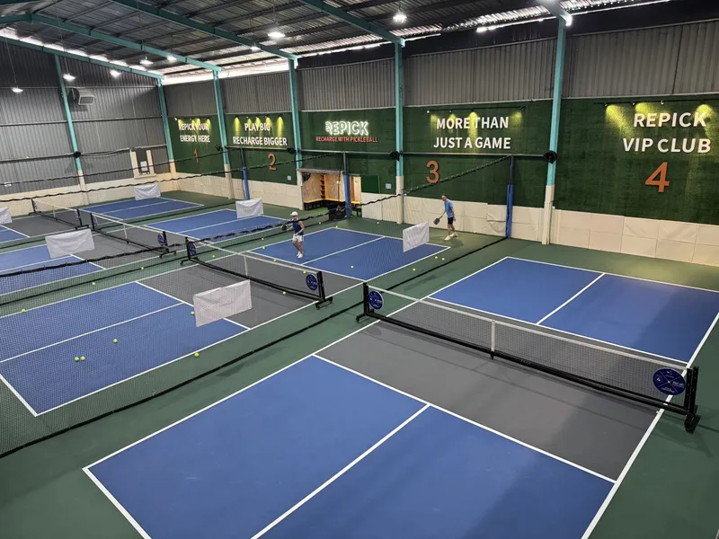 Elevated view of Repick Pickleball's indoor hall, with four blue-and-grey pickleball courts under a steel roof and green branded walls numbered 1 to 4
