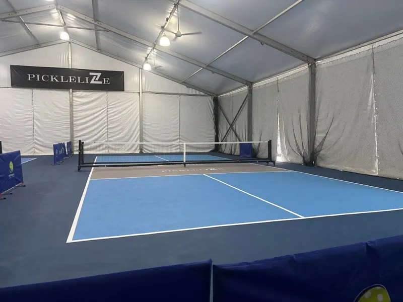 Blue pickleball court with a net under a lit white marquee tent, with a PickleliZe banner on the back wall, at PickleliZe in Pasir Ris