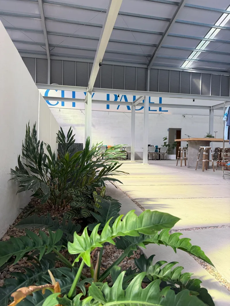 Inside City Padel KL: a roofed hall with tropical plants along a white wall, bar seating and the blue City Padel lettering on the far wall