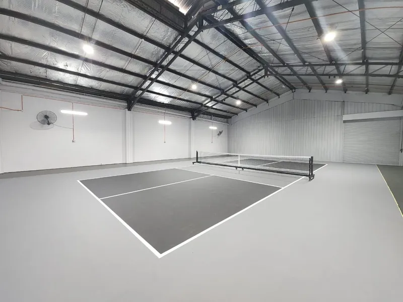 Two grey-and-charcoal indoor pickleball courts with portable nets, in a bright white warehouse hall with a steel roof truss and wall fans