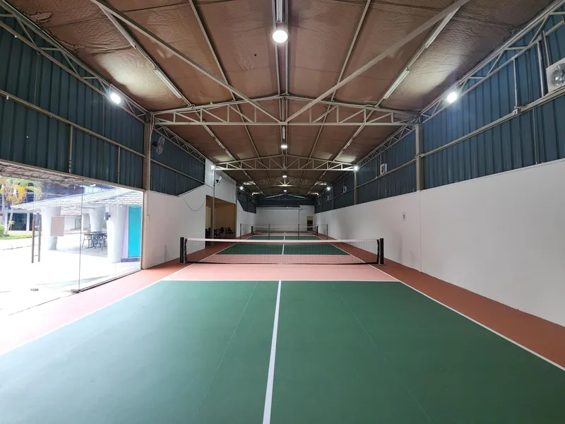 Two green pickleball courts with nets in a long indoor hall under a steel truss roof, with a glass wall opening onto the sports centre's courtyard