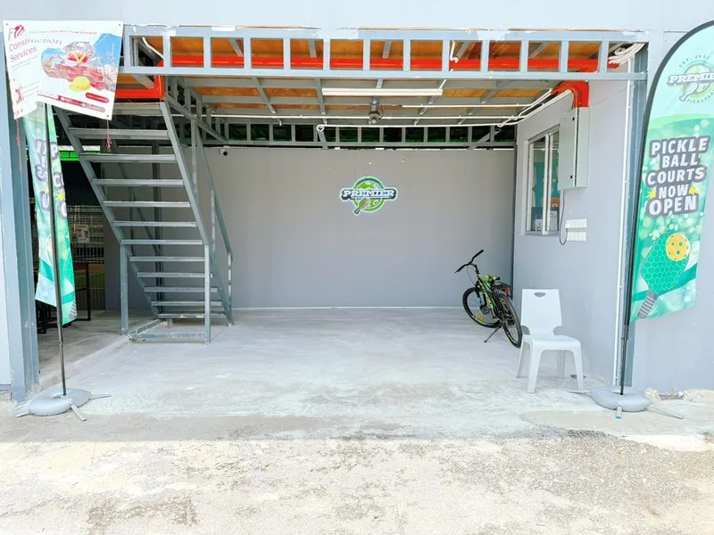 Entrance of Premier KL Pickleball, with the illuminated Premier logo on a grey wall, a steel staircase, and Premier pickleball feather flags on either side
