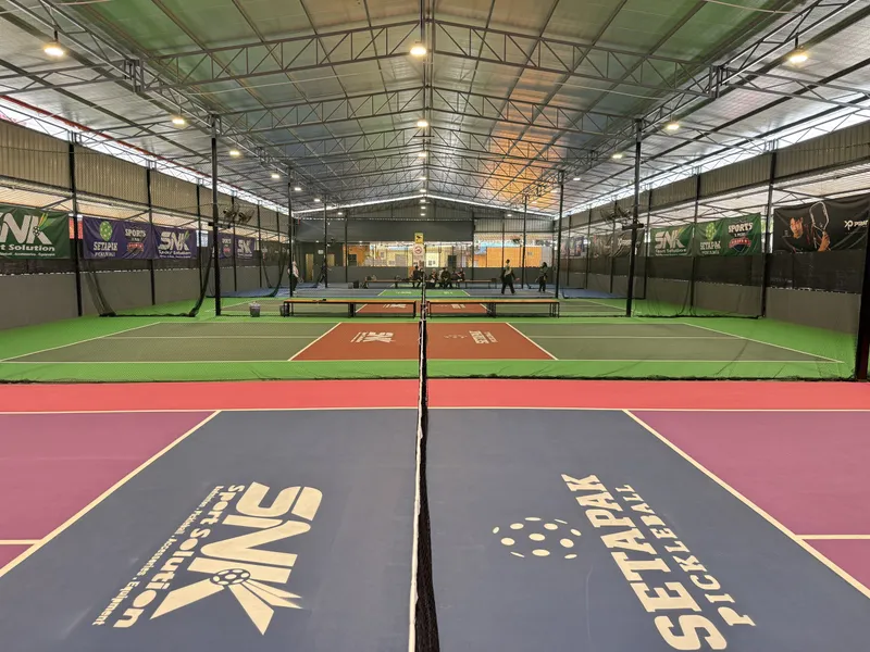 View along the net of a blue and purple pickleball court with 'Setapak Pickleball' painted on it, with more lined courts under a covered steel-roofed hall behind