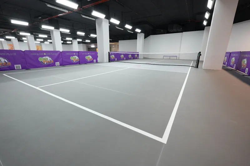 An empty indoor pickleball court with grey flooring and white lines in a bright hall, with purple One Picca barriers around the courts.