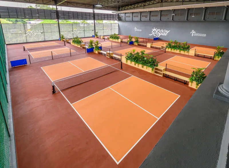 Elevated view of Pickle Royal's covered hall at Tropicana Golf & Country Resort, with orange pickleball courts, planters and benches between them, and Pickle Royal signage on the far wall