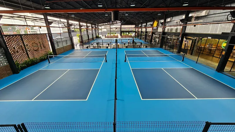 Elevated view of four blue pickleball courts with nets under a high steel roof, lined by brick screens, plants and a café