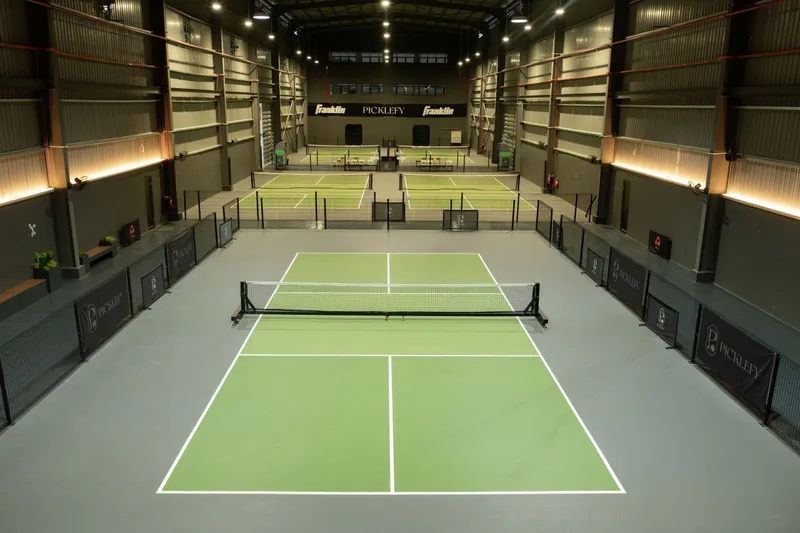 Inside Picklefy's warehouse hall: a green pickleball court with its net in the foreground, more courts behind it, Picklefy-branded fence panels and warm wall lighting