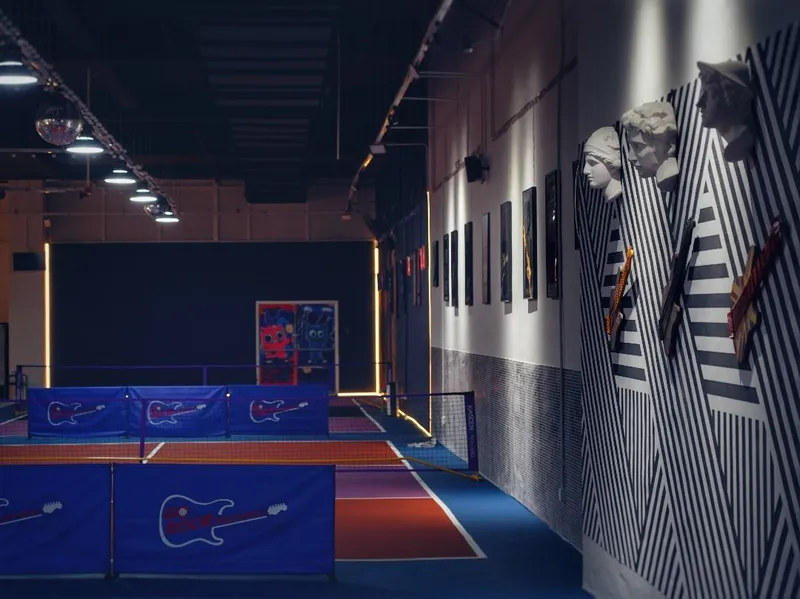 Indoor pickleball courts at Go Rock Pickleball, with blue guitar-logo net banners, red and blue court surfaces, and a rock-themed wall of striped panels, busts and guitars