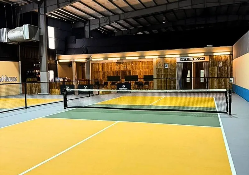 An indoor pickleball court at PickleHaus with a yellow and green surface and a net, under a high steel warehouse roof, with the reception and fitting-room area behind