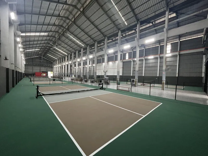 Indoor pickleball courts with brown playing surfaces and green surrounds in a large, brightly lit warehouse hall at The Backyard Pickleball, Petaling Jaya