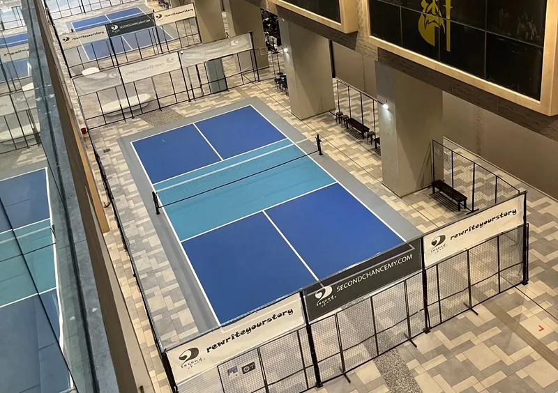 Overhead view of a blue indoor pickleball court with mesh fencing and benches in the Icon City concourse, with more courts beyond it