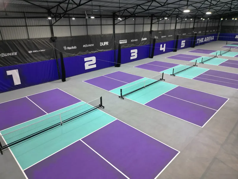 Elevated view of an indoor hall with purple and teal pickleball courts, their nets set up, and numbered courts 1 to 5 along a blue padded wall under a steel roof
