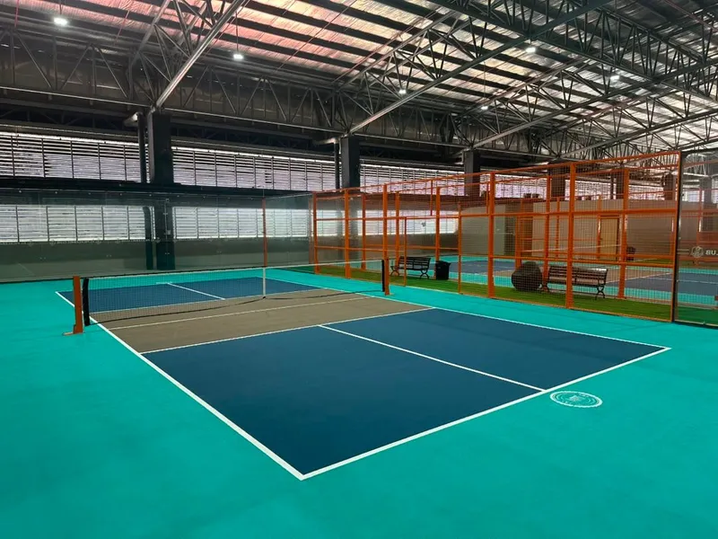 An empty indoor pickleball court with a navy playing area and teal surround under a steel-truss roof, with orange-framed caged courts and benches behind it at KL Pickleball Club, TBG Mall