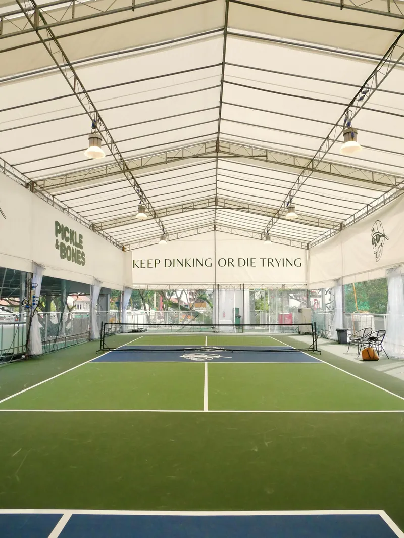 An empty pickleball court under a white sheltered tent roof at Pickle & Bones, Trifecta, with green and blue court surface and Pickle & Bones banners