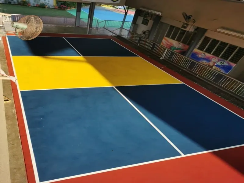 A blue and yellow pickleball court with white lines and a red border, under a covered walkway between the school's buildings, with a standing fan at one end