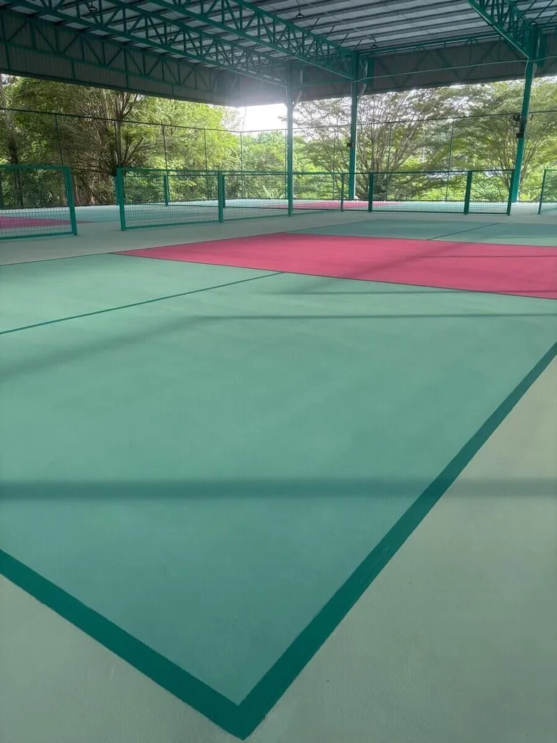 Empty green and pink pickleball courts under a steel-framed roof, with fencing between courts and trees beyond the open sides
