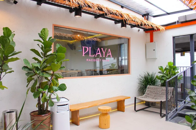 Entrance lounge of PLAYA Racquet Club at TREC KL: a white wall with a window bearing the pink PLAYA Racquet Club sign, potted plants, a wooden bench and a thatched pergola.
