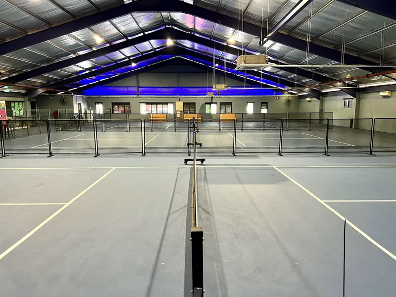 Indoor pickleball hall at Pickle Minded PJ, seen from behind a court net, with blue courts separated by low black barriers under a high steel roof