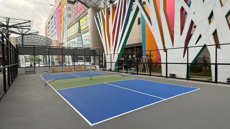 Blue and green outdoor pickleball courts behind black fencing at the main entrance of The Strand mall in Kota Damansara, under a glass canopy and beside colourful striped pillars