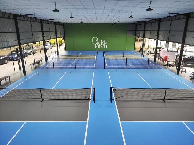 Elevated view of four blue pickleball courts with grey kitchen zones under a covered hall, with a green Pickle Porch logo wall at the far end