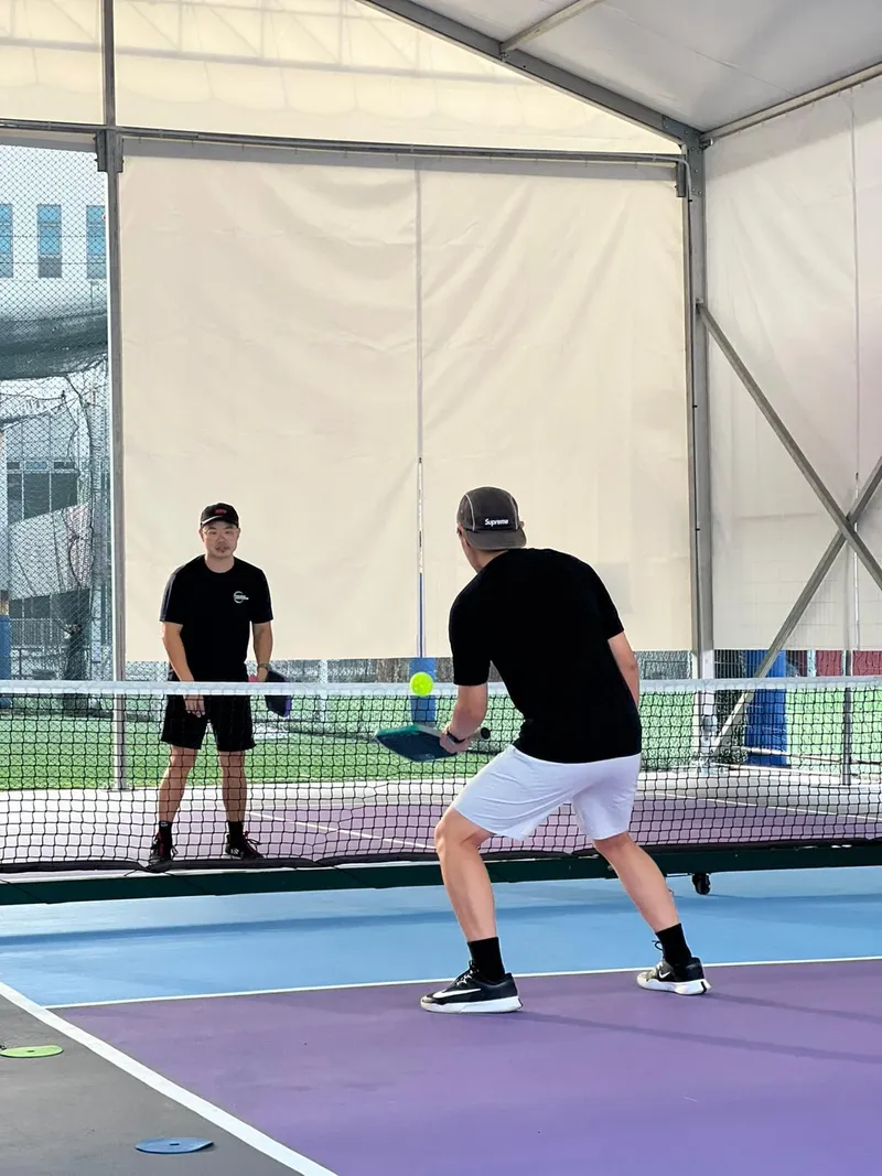 Two players rallying across the net on a purple and blue pickleball court under the covered tent at Pixel Pickle, Singapore Chinese Recreation Club