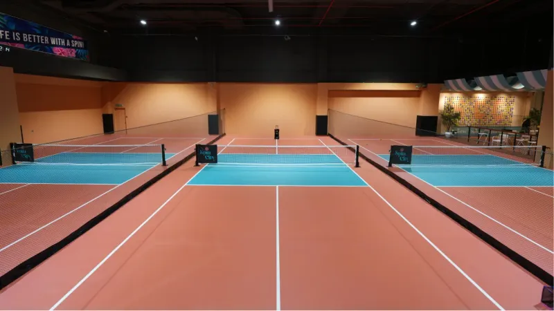 Three indoor pickleball courts at SpinCity with terracotta and teal surfaces, nets carrying SpinCity branding, and a lounge area at the back right.