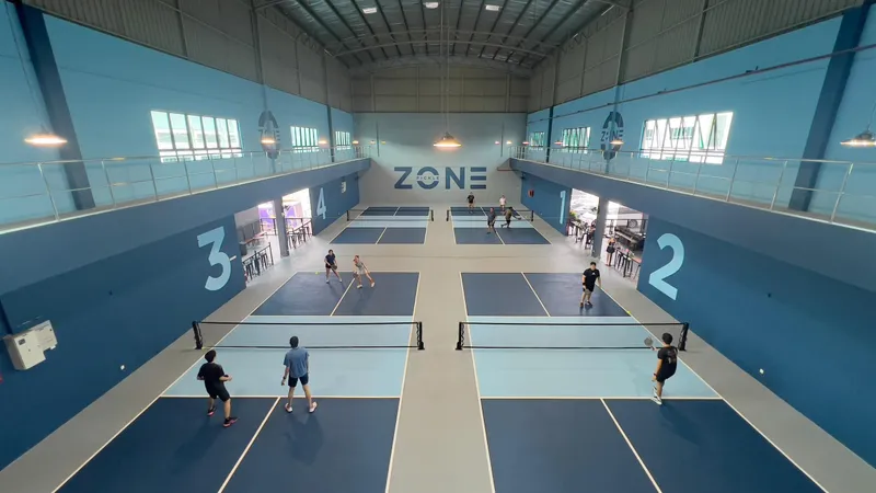 Elevated view down Pickle Zone's indoor hall: four blue numbered pickleball courts in two rows, with ZONE branding on the far wall and a mezzanine along both sides